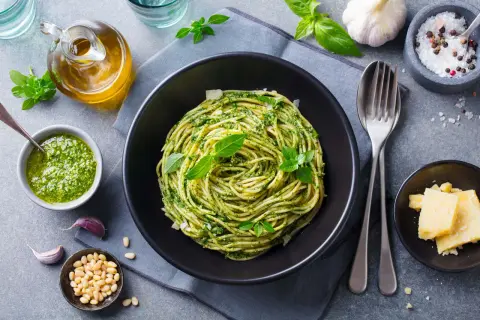 Spaghetti z pesto i bazylią w czarnej misce, obok sos, orzeszki piniowe, czosnek i parmezan.