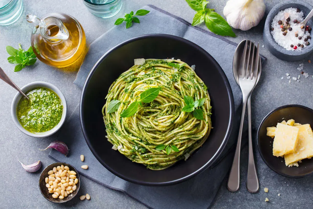 Spaghetti z pesto i bazylią w czarnej misce, obok sos, orzeszki piniowe, czosnek i parmezan.