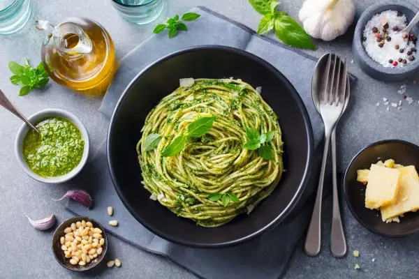 Spaghetti z pesto i bazylią w czarnej misce, obok sos, orzeszki piniowe, czosnek i parmezan.